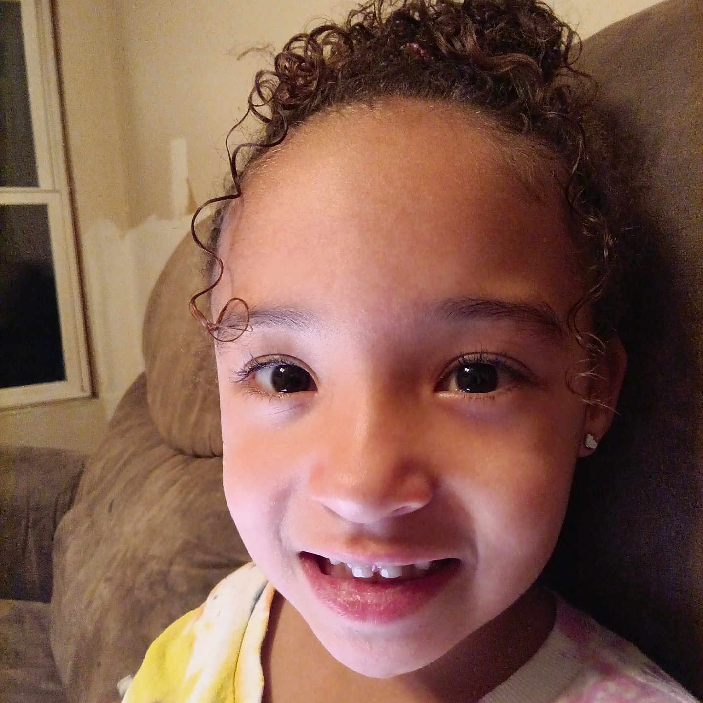 Kennedy is registered to the contest to win money with this photo: child, childhood, couch, cozy, curly_hair, earrings, eyebrows, eyelashes, face, hair, happy, indoor, living_room, person, portrait, skin, smiling, tie_dye_shirt, window, young