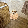 puppy, dog, towel, bathroom, floor, wicker_basket, tile_wall, pet, cute, small_dog, wood_floor, domestic, animal, indoor, resting, looking, curious, cozy, corner, home