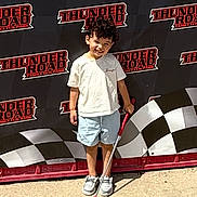 Athryn is registered to the contest to win money with this photo: child, boy, mini_golf_club, curly_hair, shorts, tshirt, sneakers, outdoor, sunny, smiling, posing, fun_park, banner, checkered_flag, concrete, recreation, happy, standing, sport, family_fun