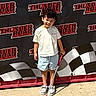 child, boy, mini_golf_club, curly_hair, shorts, tshirt, sneakers, outdoor, sunny, smiling, posing, fun_park, banner, checkered_flag, concrete, recreation, happy, standing, sport, family_fun