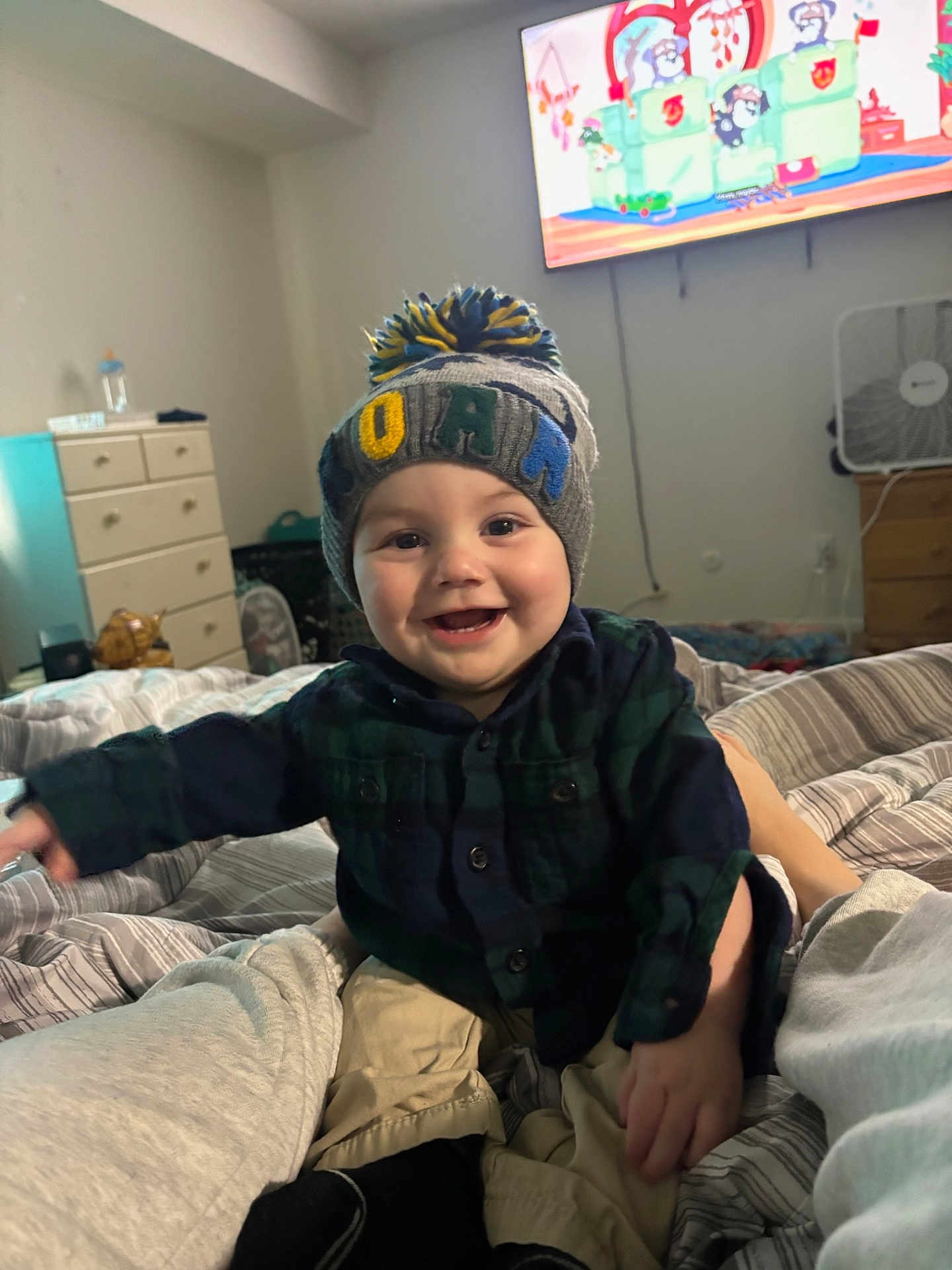 Zayden is registered to the contest to win money with this photo: baby, child, smiling, hat, knit_hat, plaid_shirt, bed, blanket, indoor, television, cartoon, dresser, fan, furniture, person, happy, cute, home, cozy, portrait