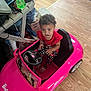 toddler, child, toy_car, barbie, indoor, curly_hair, red_clothing, polka_dot_pants, wood_floor, shoes, sneakers, plastic_toy, steering_wheel, flooring, playtime, expression, person, real_photo, casual, home