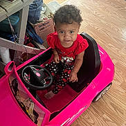 Hartley joined the competition — help win amazing prizes! toddler, child, toy_car, pink, indoor, flooring, curly_hair, clothing, red_shirt, polka_dot_pants, shoes, steering_wheel, expression, pout, seat, car_toy, floor, household_items, box, casual