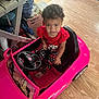 toddler, child, toy_car, pink, indoor, flooring, curly_hair, clothing, red_shirt, polka_dot_pants, shoes, steering_wheel, expression, pout, seat, car_toy, floor, household_items, box, casual
