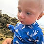 baby, child, close_up, blue_clothing, beach, sand, outdoor, curious, bald_head, patterned_shirt, sea, summer, vacation, sunny, young_child, sitting, casual, daytime, nature, portrait