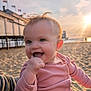 baby, child, smiling, pink_clothing, beach, sand, sunset, pier, flags, happy, toddler, portrait, cute, outdoor, sunlight, hand_in_mouth, warm_light, hair, face, ocean