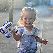 Avalynn is registered to the contest to win money with this photo: child, toddler, bubble_gun, bubbles, smiling, happy, outdoor, playful, overalls, blonde_hair, sunlight, face, person, toy, fun, summer, casual_clothing, young_child, joy, portrait
