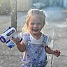 child, toddler, bubble_gun, bubbles, smiling, happy, outdoor, playful, overalls, blonde_hair, sunlight, face, person, toy, fun, summer, casual_clothing, young_child, joy, portrait