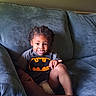 child, toddler, boy, smiling, portrait, indoors, couch, sitting, batman, superhero, costume, t_shirt, braids, curly_hair, barefoot, legs, hand, living_room, upholstery, happy