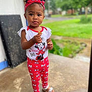 Jah'Liya joined the competition — help win amazing prizes! child, toddler, girl, portrait, standing, outdoor, doorway, grass, trees, red_headband, curly_hair, smiling, face, snack, food, minnie_mouse, disney, patterned_clothing, sandals, daylight