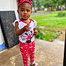 child, toddler, girl, portrait, standing, outdoor, doorway, grass, trees, red_headband, curly_hair, smiling, face, snack, food, minnie_mouse, disney, patterned_clothing, sandals, daylight