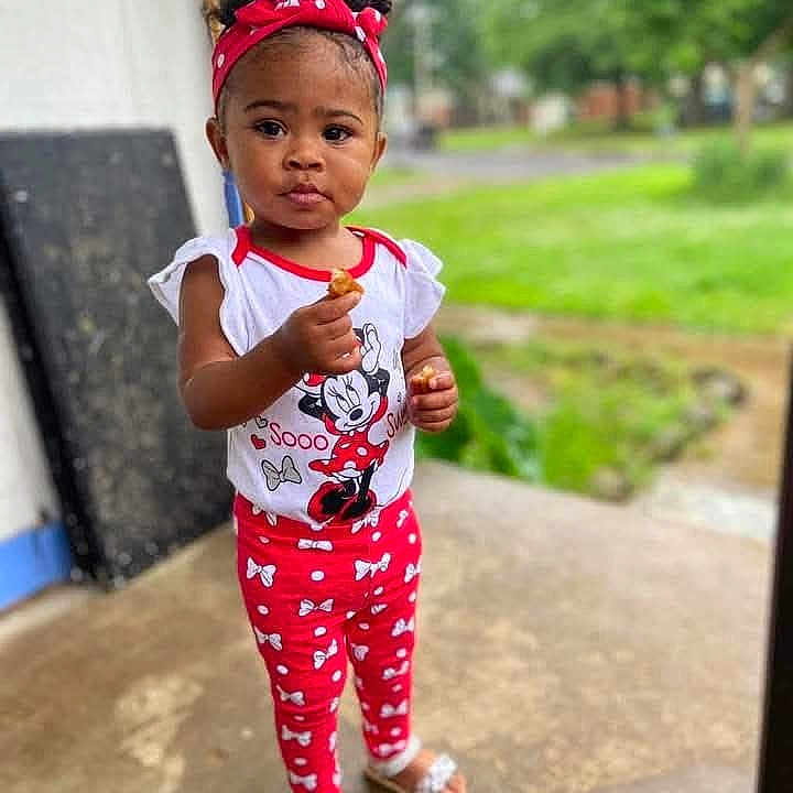 Jah'Liya joined the competition — help win amazing prizes! child, toddler, girl, portrait, standing, outdoor, doorway, grass, trees, red_headband, curly_hair, smiling, face, snack, food, minnie_mouse, disney, patterned_clothing, sandals, daylight