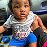 toddler, child, baby, portrait, sitting, car, indoors, curly_hair, dark_skin, wide_eyes, facial_expression, gray_shirt, blue_shorts, adult, arm, close_up, automotive_interior, seated, daylight, photography