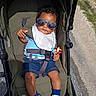 baby, child, stroller, sunglasses, outdoor, portrait, seated, smiling, toy, bib, shirt, shorts, socks, sneakers, harness, infant, pavement, grass, sunlight, fashion