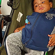 Tatum joined the competition — help win amazing prizes! baby, child, infant, stroller, portrait, face, serious_expression, curly_hair, blue_hoodie, gray_clothing, seated, indoor, close_up, baby_carriage, fabric, zipper, hand, legs, photography, safe