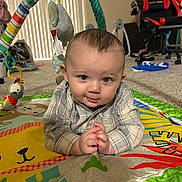 Malachi joined the competition — help win amazing prizes! baby, infant, play_mat, toy, indoor, child, crawling, colorful, cute, person, face, hands, plaid_shirt, carpet, furniture, gaming_chair, soft_lighting, curious, eyes, expression