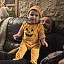 toddler, man, pumpkin_costume, couch, cat, window, stone_wall, indoor, smile, child, adult, furniture, home, beard, black_clothing, orange_outfit, hat, pet, cozy, relaxation