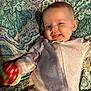 baby, infant, child, smiling, gummy_smile, onesie, unicorn_print, rainbow_print, rattle, toy, hand, cheek, tongue, eyes, pajamas, blanket, patterned_bedspread, pillow, lying_down, portrait