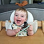 Léon participe au concours pour gagner de l'argent avec cette photo : baby, high_chair, spoon, bib, smiling, happy, indoor, child, furniture, table, wood, face, person, cheerful, young_child, living_room, couch, fireplace, clothing, playful