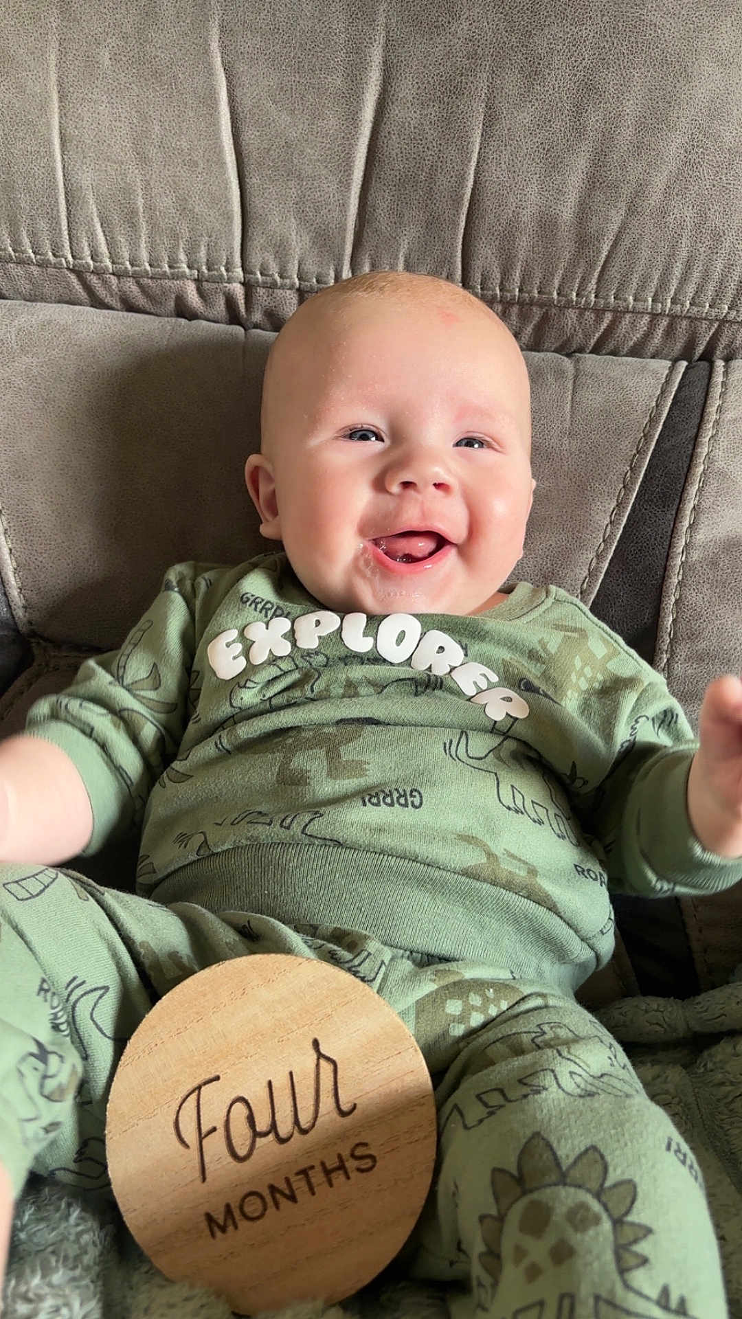 Harley is registered to the contest to win money with this photo: baby, infant, smiling, green_clothing, dinosaur_print, wooden_sign, four_months, sitting, soft_chair, cozy, happy, cute, milestone, portrait, child, indoors, comfortable, young_baby, cute_expression, playful