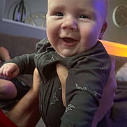 Harley is registered to the contest to win money with this photo: baby, smiling, onesie, hand, living_room, person, infant, face, child, indoor, happy, holding, couch, furniture, adult, soft_lighting, portrait, closeup, cute, expression