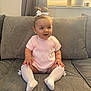 Remy is registered to the contest to win money with this photo: baby, child, sitting, pink_dress, white_socks, bow, hair_tie, couch, gray_cushions, indoor, happy, smiling, cute, childrens_clothing, legs, hands, window, curtains, home, portrait