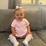 Remy is registered to the contest to win money with this photo: baby, child, pink_outfit, white_bow, sitting, couch, gray_couch, white_socks, knee_high_socks, indoors, portrait, cute, smiling, blue_eyes, natural_light, home_interior, window, curtains, bracelet, plush