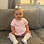 baby, child, pink_outfit, white_bow, sitting, couch, gray_couch, white_socks, knee_high_socks, indoors, portrait, cute, smiling, blue_eyes, natural_light, home_interior, window, curtains, bracelet, plush