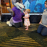 Savannah is registered to the contest to win money with this photo: toddler, child, mascot, wolf, costume, animal, meeting, greeting, indoor, event, staff_member, dress, colorful_clothing, barefoot, smiling, carpet, stage, backdrop, family_friendly, woodland_theme