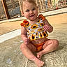 Savannah is registered to the contest to win money with this photo: baby, child, infant, smiling, portrait, swimming_pool, water, outdoor, sunlight, floral_dress, sunflowers, orange_clothing, sitting, wet, play_area, fence, playground, summer, barefoot, happy