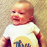 Gunnar is registered to the contest to win money with this photo: baby, smiling, infant, onesie, carpet, lying_down, child, cute, happy, face, clothing, celebration, milestone, skin, person, indoors, young_child, cheerful, head, expression