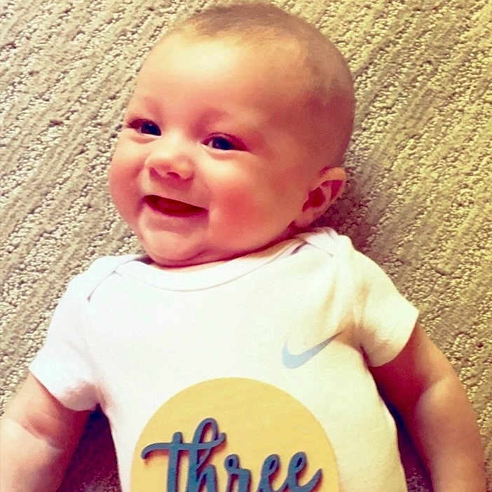 Gunnar is registered to the contest to win money with this photo: baby, smiling, infant, onesie, carpet, lying_down, child, cute, happy, face, clothing, celebration, milestone, skin, person, indoors, young_child, cheerful, head, expression