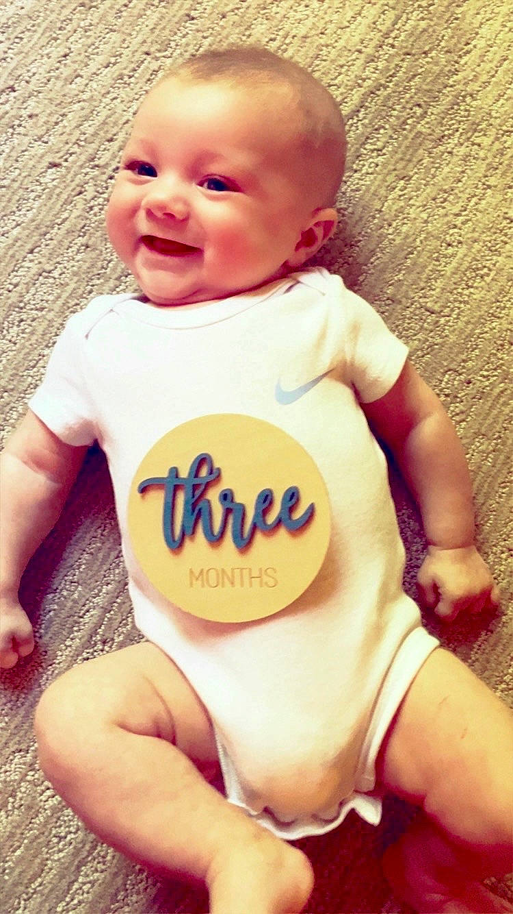 Gunnar is registered to the contest to win money with this photo: baby, carpet, cheerful, child, clothing, cute, face, happy, human, indoors, infant, lying_down, onesie, person, skin, small, smiling, sticker, three_months, young