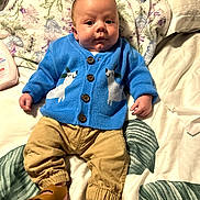Cayden is registered to the contest to win money with this photo: baby, infant, child, blue_sweater, polar_bear_design, tan_pants, brown_sandals, blanket, leaf_pattern, bed, lying_down, cute, curious_expression, indoors, soft_lighting, portrait, clothing, footwear, person, closeup