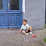 Yassine a rejoint le concours — aidez-le/la à gagner de superbes lots ! child, boy, curly_hair, sitting, street, cobblestone, blue_door, gray_wall, sidewalk, casual, outdoor, daylight, clothing, shorts, sneakers, red_cloth, urban, young, thoughtful, curious