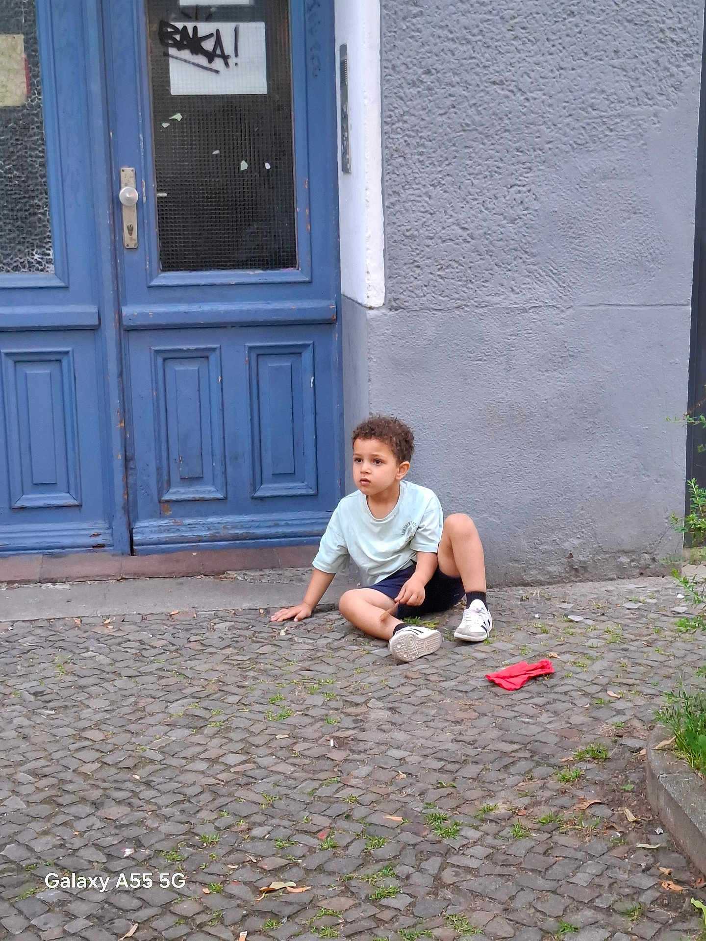Yassine a rejoint le concours — aidez-le/la à gagner de superbes lots ! child, boy, curly_hair, sitting, street, cobblestone, blue_door, gray_wall, sidewalk, casual, outdoor, daylight, clothing, shorts, sneakers, red_cloth, urban, young, thoughtful, curious