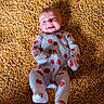 baby, infant, child, smiling, happy, lying_down, portrait, face, eyes, mouth, onesie, clothing, pumpkin_pattern, red_pattern, blanket, textured_background, golden, corn_kernels, indoor, close_up