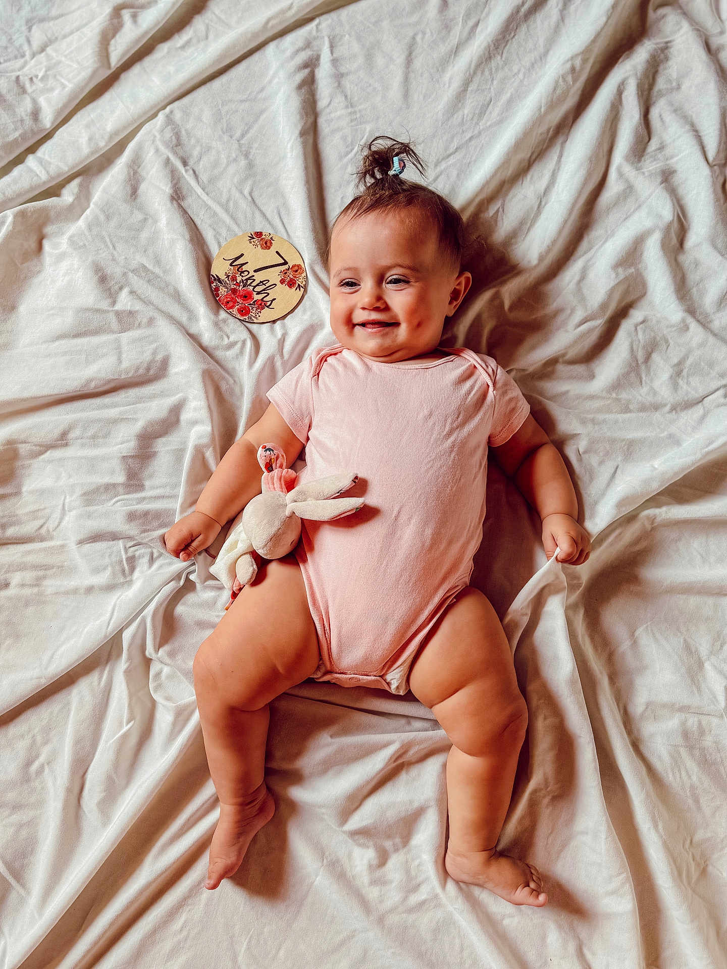 Alma a rejoint le concours — aidez-le/la à gagner de superbes lots ! baby, infant, pink_onesie, smiling, dimples, plush_toy, bunny, pony_tail, white_sheet, milestone_card, 7_months, lying_down, happy, cute, child, skin, hand, foot, portrait, indoors