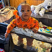Kingston is registered to the contest to win money with this photo: baby, child, playpen, toy, stuffed_animal, orange_sweater, indoor, wood_paneling, curious, standing, infant, face, hand, floor, plush_toy, colorful_toys, closeup, expression, home, cute