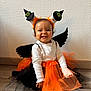 child, costume, cute, festive, floor, halloween, happy, headband, holiday, indoor, orange_tutu, playful, portrait, sitting, smiling, toddler, wall, white_shirt, wings, witch_hat