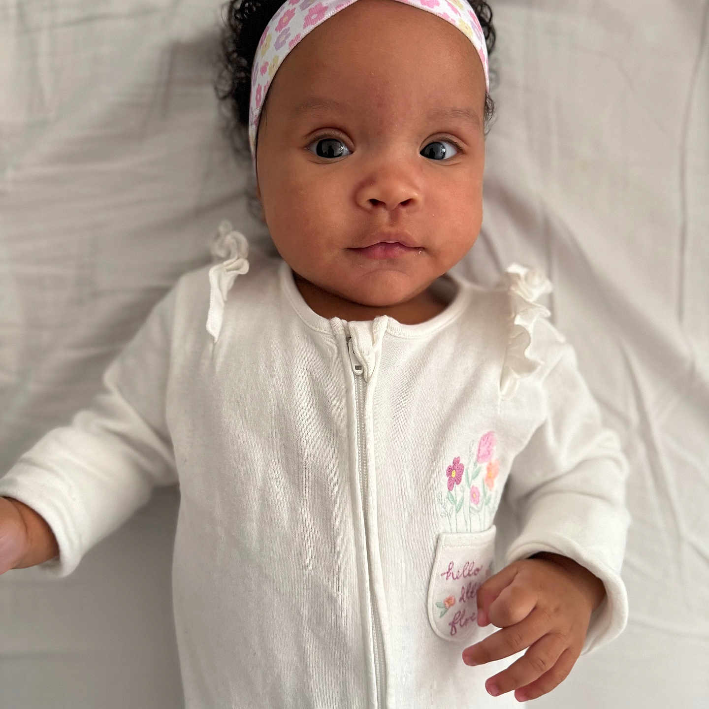 Teyana participe au concours pour gagner de l'argent avec cette photo : baby, blanket, child, close_up, curly_hair, cute, expression, face, hand, headband, indoor, infant, looking_at_camera, lying_down, onesie, person, portrait, soft_light, white_clothing, young_child