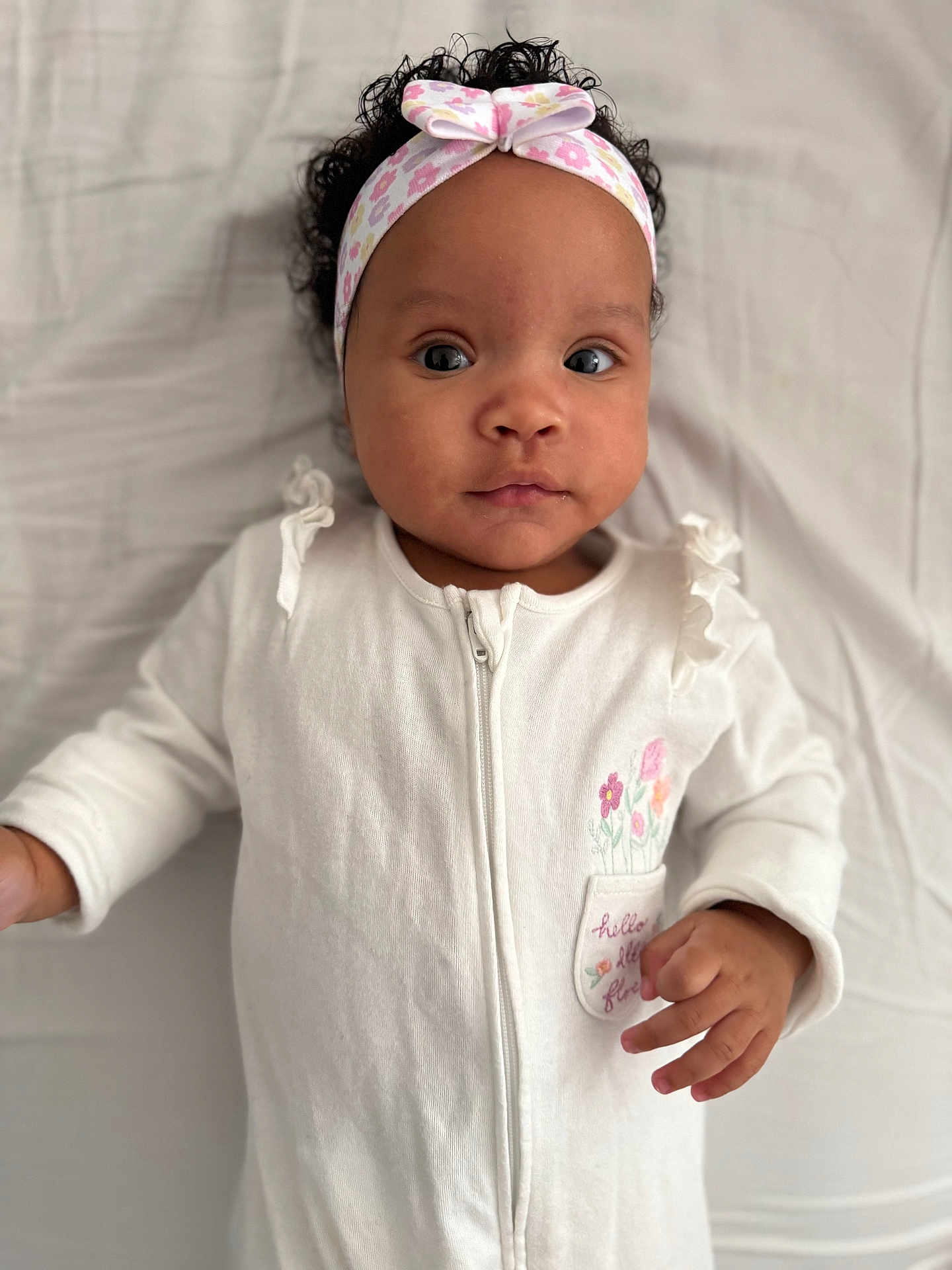 Teyana participe au concours pour gagner de l'argent avec cette photo : baby, infant, headband, white_clothing, onesie, curly_hair, lying_down, blanket, portrait, close_up, child, cute, indoor, young_child, person, face, hand, looking_at_camera, soft_light, expression