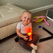 Danial is registered to the contest to win money with this photo: toddler, child, smiling, indoor, carpet, toy, balloon, ottoman, white_shirt, pants, socks, floor, home, play, colorful, light, glowing, happy, cute, person