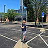 asphalt, blue_sky, boy, casual_clothing, child, concrete_base, daytime, hand_holding, nature, outdoor, parking_lot, person, playful, pole, shorts, smile, sneakers, standing, sunny, tree