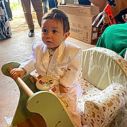 Gabriel participe au concours pour gagner de l'argent avec cette photo : toddler, child, white_suit, toy, wooden_toy, indoor, chair, embroidered, people, adults, party, celebration, curious, decor, box, casual_clothing, floor, person, footwear, green