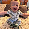 arms_outstretched, baby, bed, bedroom, child, cozy, cute, dinosaur_pajamas, footwear, happy, indoors, infant, patterned_bedspread, person, pillow, plush_slippers, sitting, sleepwear, smiling, wooden_headboard
