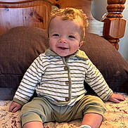 Wyatt is registered to the contest to win money with this photo: baby, child, smile, curly_hair, striped_jacket, green_pants, socks, bed, pillow, wooden_headboard, indoor, happy, cute, person, infant, portrait, seated, cozy, comfortable, home