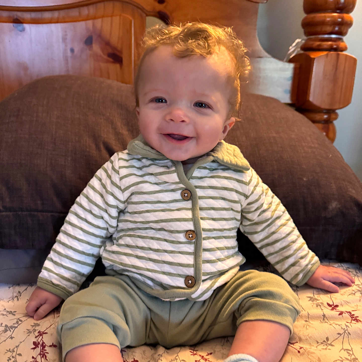Wyatt is registered to the contest to win money with this photo: baby, child, smile, curly_hair, striped_jacket, green_pants, socks, bed, pillow, wooden_headboard, indoor, happy, cute, person, infant, portrait, seated, cozy, comfortable, home