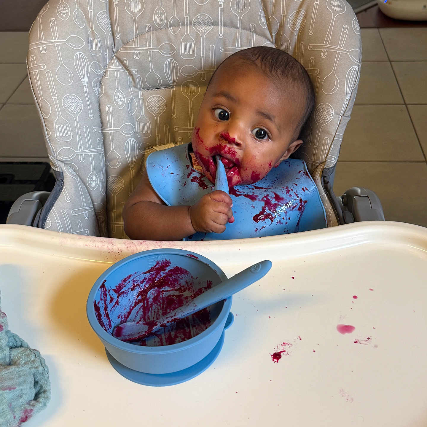 Shiloh joined the competition — help win amazing prizes! baby, high_chair, bib, spoon, bowl, food_stains, messy, curious, indoor, kitchen, child, feeding, purple_food, table, seat, utensils_pattern, cloth, cute, expression, toddler