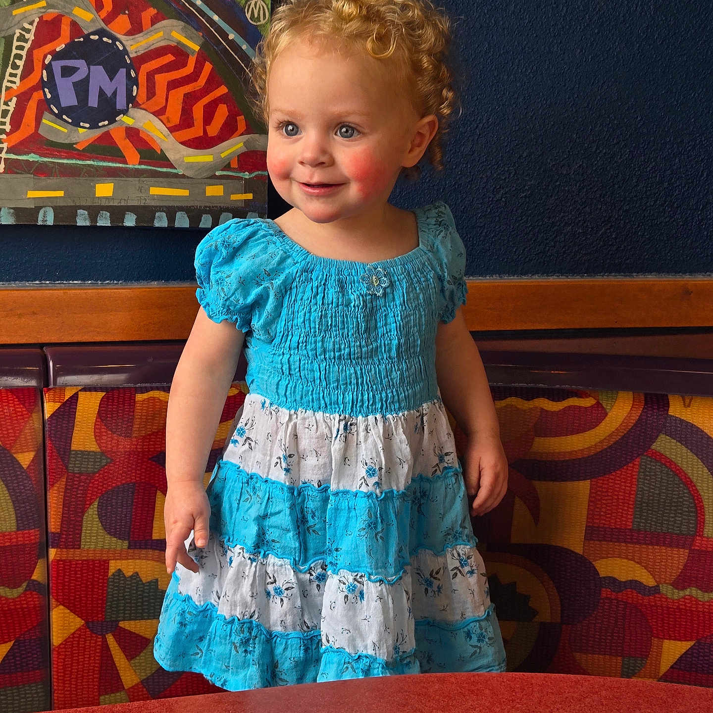 Nora joined the competition — help win amazing prizes! bench, blue_dress, child, clothing, colorful, curly_hair, cute, face, happy, indoor, light, patterned_furniture, person, portrait, rosy_cheeks, smiling, standing, table, toddler, wall_art